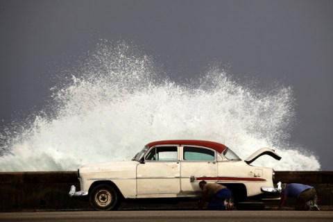 Men change a flat tyre of their car on Havana's seafront boulevard as a wave crashes
