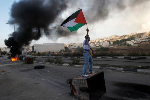 Palestinian stone-thrower holds a flag as he stands atop a garbage bin during clashes with Israeli security forces near Ramallah