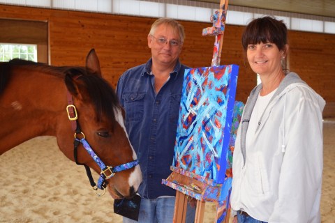 Metro, a 10-year-old retired bay thoroughbred horse stands with owners Ron Krajewski and Wendy Krajewski (R) and one of his paintings at Motter's Station Stables in Rocky Ridge