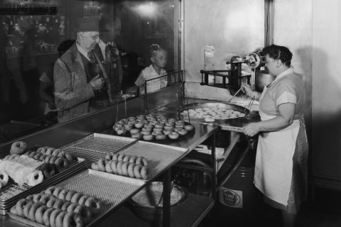 A baker checks the progress of the donuts she is frying as customers watch the donuts being prepared through a window.