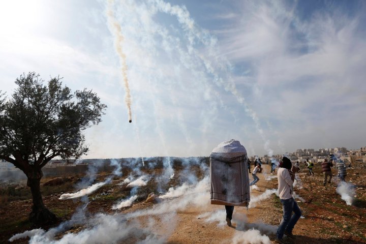 A man wearing a large puppet head and Palestinian stone-throwing protesters run as tear gas is fired by Israeli soldiers during clashes at a weekly demonstration against Jewish settlements in the West Bank village of Bilin, near Ramallah Jan. 3, 2014. 